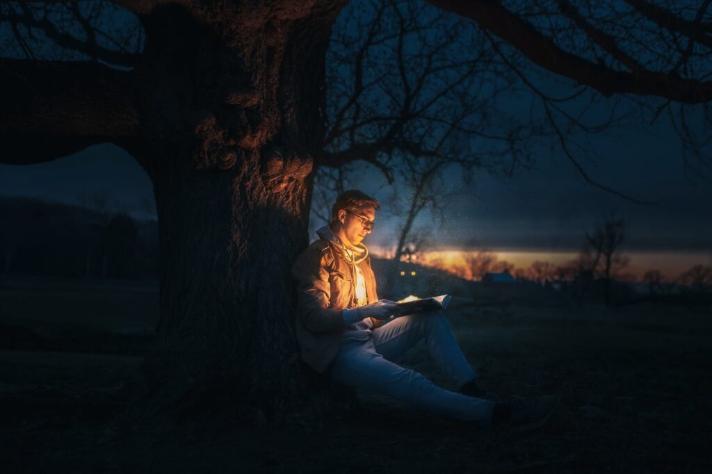 man sitting under a tree reading a book during night time. World Book Night 2023