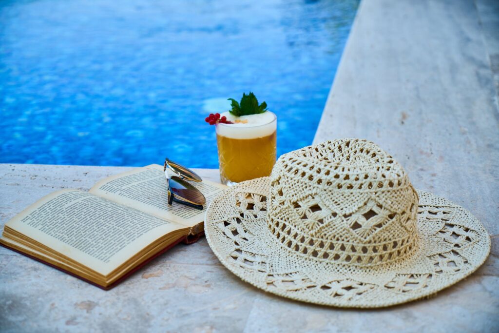 beige straw hat book sunglasses and drink beside pool. Best books in June 2023: recommendations
