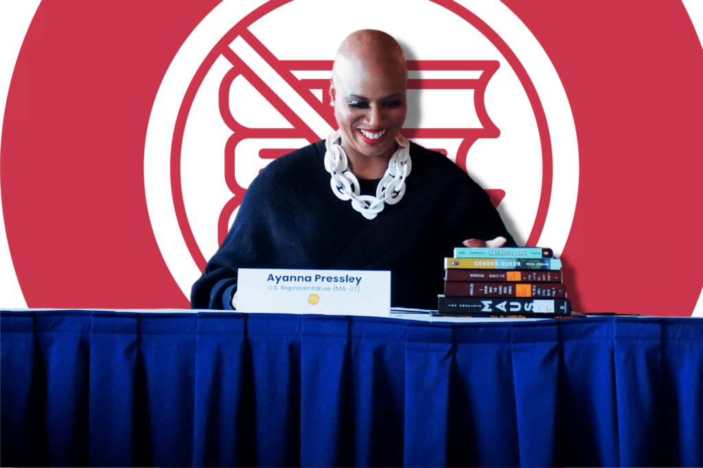 Rep Ayanna Pressley introduces Books Save Lives Act, sitting at a table with a blue table cloth, with a stack of banned books, a sign with her name on the table and a red and white logo of banned books