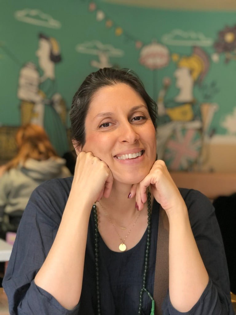 Nazanin Zaghari-Ratcliffe talks about the power of books during her time in an Iranian prison. She is seen wearing a green beaded necklace and a grey long-sleeved top, while smiling at the camera and leaning on arms. Green motif in the background.