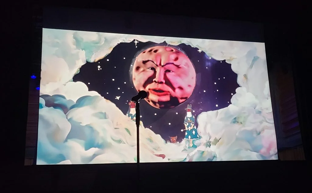The image shows a stage with a microphone stand in front of a large screen displaying an animated graphic. The graphic features a stylized image of the moon with a human face, surrounded by clouds and stars. The moon's expression is calm, with eyes closed, set against a dark night sky. Below the moon, small figures appear on clouds, including one holding a lantern, enhancing a dreamlike, whimsical atmosphere for Rough Trade Books event