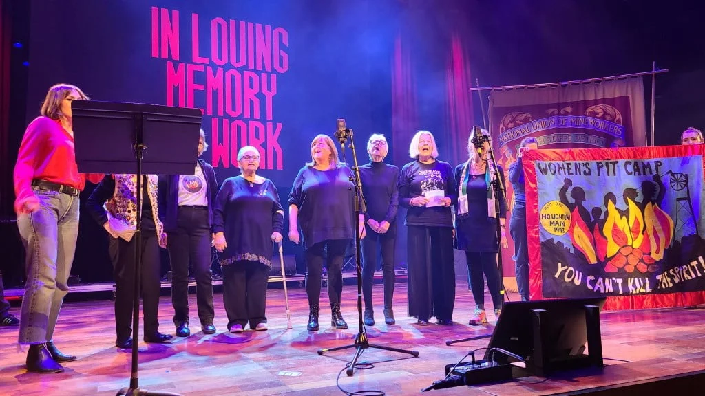 The image features a group of women on stage at an event, performing or preparing to perform. Behind them, a large banner displays the text "IN LOVING MEMORY OF WORK" in bold red letters on a blue background. To the right, two banners are held aloft; one reads "National Union of Mineworkers (South Staffs District)," and the other, more colorful banner says "Women's Pit Camp" with a motif of women around a fire and the phrase "You can't kill the spirit." The women on stage are dressed casually, some in dark colors, with one holding a microphone. The overall scene suggests a powerful, commemorative atmosphere, likely relating to historical labour movements or a specific workers' strike.